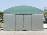 A green-domed canvas warehouse stands under a clear sky, surrounded by sparse greenery